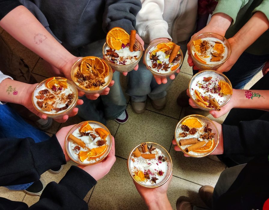 Grupa dzieci trzymająca w dłoniach własnoręcznie wykonane świece sojowe w szklanych naczyniach. Świece są bogato zdobione plastrami suszonej pomarańczy, laskami cynamonu, anyżem i szyszkami. Na przedramionach dzieci widoczne kolorowe tatuaże brokatowe