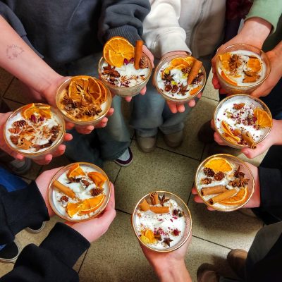 Grupa dzieci trzymająca w dłoniach własnoręcznie wykonane świece sojowe w szklanych naczyniach. Świece są bogato zdobione plastrami suszonej pomarańczy, laskami cynamonu, anyżem i szyszkami. Na przedramionach dzieci widoczne kolorowe tatuaże brokatowe