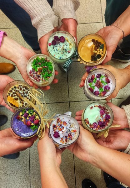 Grupa uczestników trzymająca w dłoniach własnoręcznie wykonane świece sojowe w szklanych naczyniach. Każda świeca jest inna: w kolorach fioletu, zieleni i beżu, zdobiona suszonymi kwiatami, mchem, ziarnami kawy oraz rafią