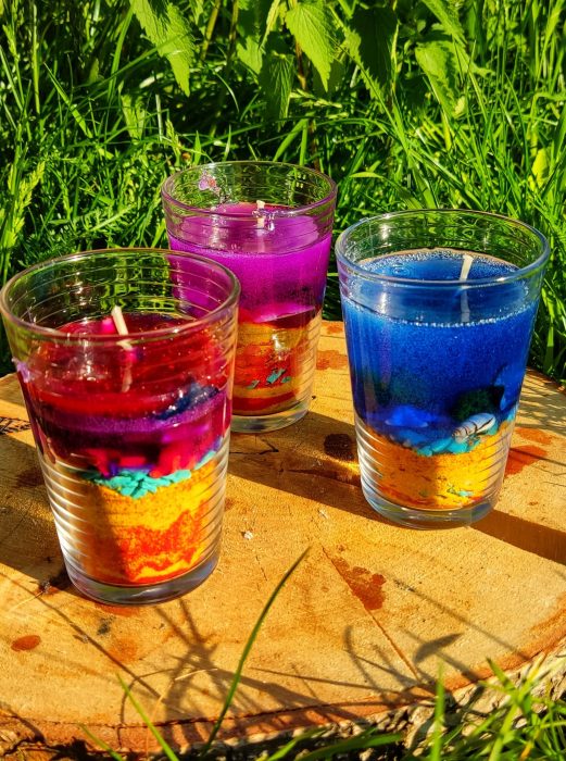 Trzy kolorowe świece żelowe w szklankach: czerwono-fioletowa, fioletowa i niebieska. Świece mają warstwową strukturę z kolorowym piaskiem i muszelkami na dnie, ustawione na drewnianym pniu w pełnym słońcu