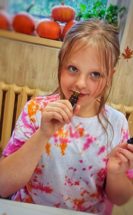 Dziewczynka w kolorowej koszulce tye-dye z uśmiechem wącha olejek eteryczny z małej brązowej buteleczki. W tle widoczne jesienne dekoracje z dyń na parapecie