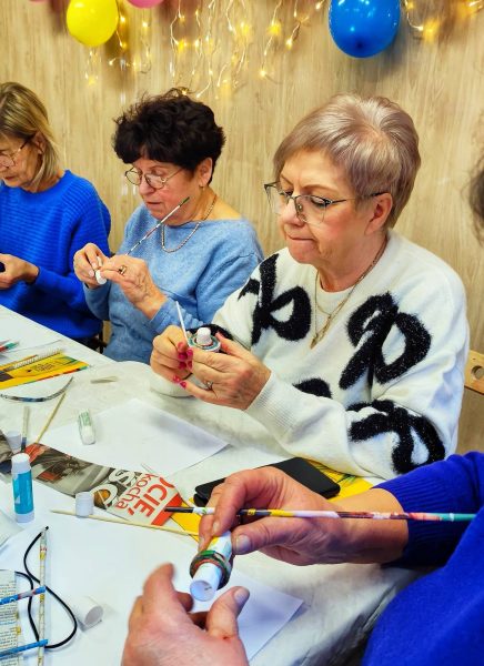 Trzy kobiety siedzą przy stole warsztatowym przykrytym białym obrusem, intensywnie pracując nad elementami z wikliny papierowej. Na pierwszym planie uczestniczka w białym swetrze z czarnymi kokardami precyzyjnie skręca kolorowy papier wokół małego, białego pojemnika. Obok niej kolejna kobieta w błękitnym swetrze smaruje papierową rurkę klejem, a w dolnej części zdjęcia widoczne są dłonie instruktorki lub innej uczestniczki pomagającej przy pracy. Na stole leżą tubki kleju, nożyczki oraz kolorowe gazety