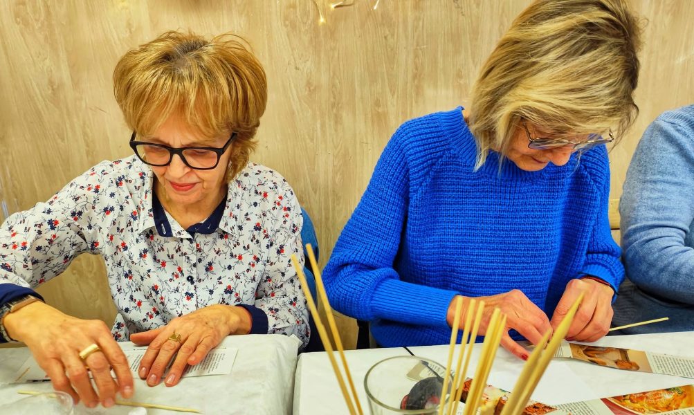 Dwie kobiety siedzą obok siebie przy stole warsztatowym, intensywnie pracując nad elementami z wikliny papierowej. Uczestniczka po lewej, w kwiecistej koszuli i okularach, precyzyjnie wygładza papierową rurkę na stole. Kobieta po prawej, w niebieskim swetrze, jest skupiona na zaginaniu papieru wokół cienkiego patyczka. Na pierwszym planie widoczne są wysokie szklanki z licznymi, jasnymi rurkami przygotowanymi do wyplatania