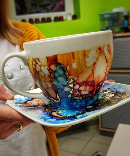Biała ceramiczna filiżanka ze spodkiem, trzymana w dłoniach, udekorowana intensywnymi tuszami alkoholowymi w kolorach błękitu, pomarańczu, brązu i fioletu. Wzór tworzy gęste, organiczne plamy z wyraźnymi złotymi akcentami szlagmetalu na krawędziach kolorów. Osoba trzymająca naczynie ma na sobie musztardowy sweter i biały fartuch