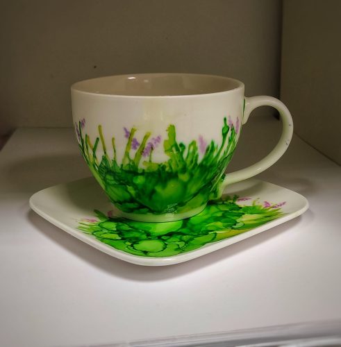 Ceramiczna biała filiżanka i kwadratowy spodek udekorowane tuszami alkoholowymi w kolorach soczystej zieleni i jasnego fioletu. Wzór w dolnej części naczynia przypomina gęstą trawę z drobnymi fioletowymi kwiatami, wykonany techniką rozproszonych plam. Zestaw ustawiony jest w białej wnęce