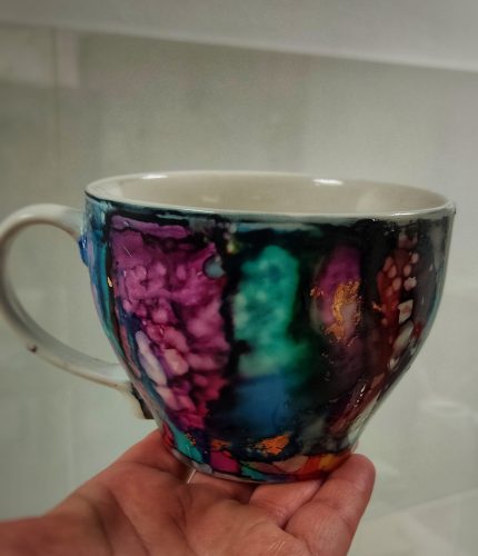 Filiżanka ceramiczna z białą rączką trzymana w dłoni, ozdobiona pionowymi pasami przenikających się barw tuszu alkoholowego w odcieniach fioletu, amarantu, szmaragdowej zieleni i granatu. Wzór posiada liczne, nieregularne złocenia szlagmetalem, które podkreślają strukturę kolorystyczną naczynia