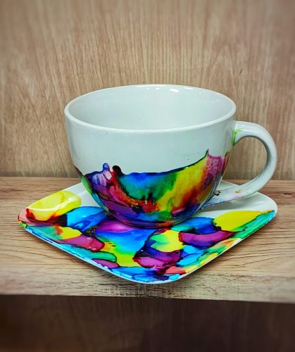 Biała ceramiczna filiżanka i kwadratowy spodek udekorowane płynnymi, przenikającymi się plamami tuszu alkoholowego w kolorach intensywnego błękitu, zieleni, fioletu, żółci i czerwieni. Wzór na naczyniach ma czyste, nasycone barwy o miękkich przejściach tonalnych. Zestaw stoi na jasnej, drewnianej półce