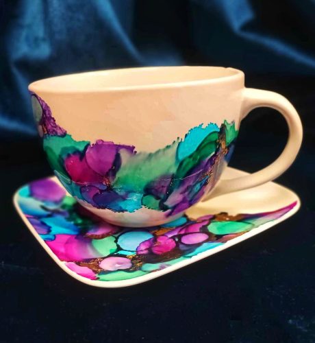 Biała ceramiczna filiżanka ze spodkiem, ozdobiona tuszami alkoholowymi w odcieniach głębokiego fioletu, turkusu i morskiej zieleni. Wzór przypomina nakładające się na siebie płatki kwiatów lub morskie fale, z licznymi, drobnymi akcentami złotego szlagmetalu na krawędziach kolorów. Zestaw wyeksponowany na ciemnym, granatowym tle