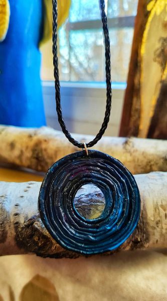 Okrągły wisior z otworem w środku, wykonany z ciasno plecionej wikliny papierowej o wyraźnej, spiralnej strukturze. Biżuteria jest pomalowana na ciemnogranatowy, niemal czarny kolor z połyskiem, przez który przebijają subtelne, różowe refleksy. Wisior zawieszony jest na plecionym, czarnym rzemyku i spoczywa na pniach brzozy o jasnej korze