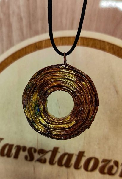 Okrągły wisior z otworem w środku, wykonany z ciasno zwiniętej wikliny papierowej o wyraźnej, warstwowej strukturze. Biżuteria jest pomalowana na metaliczny, złoty kolor z widocznymi przetarciami w odcieniach miedzi, zieleni i błękitu. Wisior wisi na cienkim, czarnym rzemyku na tle jasnego drewna z fragmentem napisu "Warsztatownia"