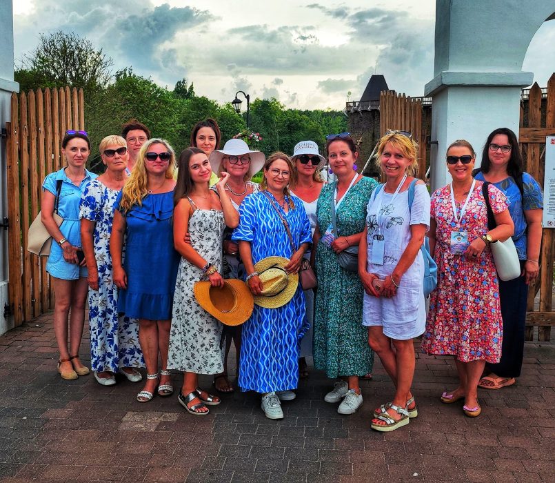 Grupa uśmiechniętych kobiet w letnich sukienkach i kapeluszach pozuje do wspólnego zdjęcia przed drewnianą bramą w Inowrocławiu. W środku grupy Monika Wojdyło w zielonej sukience i okularach przeciwsłonecznych