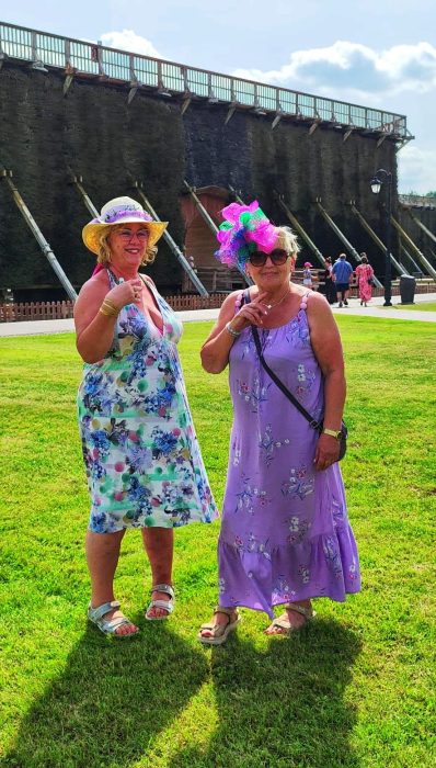 Dwie uśmiechnięte kobiety w letnich sukienkach i strojnych kapeluszach z kwiatami pozują na trawiastym terenie przed tężnią solankową w Inowrocławiu