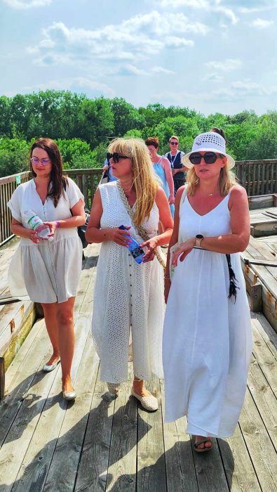 Trzy kobiety w letnich, białych i jasnych sukienkach spacerują po nasłonecznionym, drewnianym pomoście tężni solankowej w Inowrocławiu