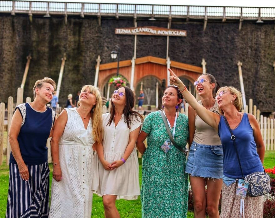Sześć uśmiechniętych kobiet pozuje przed tężnią solankową w Inowrocławiu; jedna z nich w niebieskiej koszulce wskazuje ręką w górę, a Monika Wojdyło w zielonej sukience patrzy w tym samym kierunku