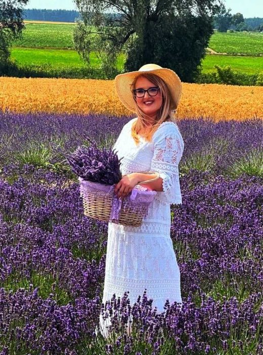 Kobieta w białej, koronkowej sukience i słomkowym kapeluszu stoi na polu lawendy, trzymając wiklinowy kosz po brzegi wypełniony świeżo ściętymi, fioletowymi kwiatami