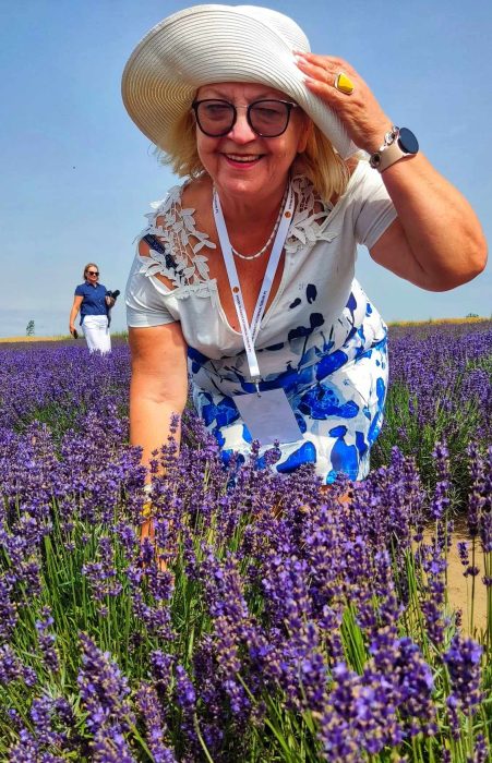 Uśmiechnięta kobieta w białym kapeluszu przeciwsłonecznym i biało-niebieskiej sukience pochyla się nad rzędami kwitnącej, fioletowej lawendy na polu pod błękitnym niebem