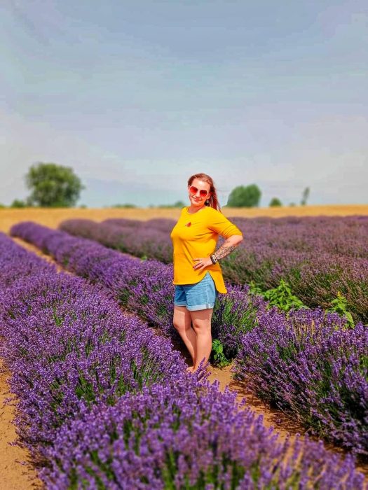 Kobieta w żółtej bluzce i okularach przeciwsłonecznych stoi uśmiechnięta pośród równoległych rzędów kwitnącej lawendy pod błękitnym niebem