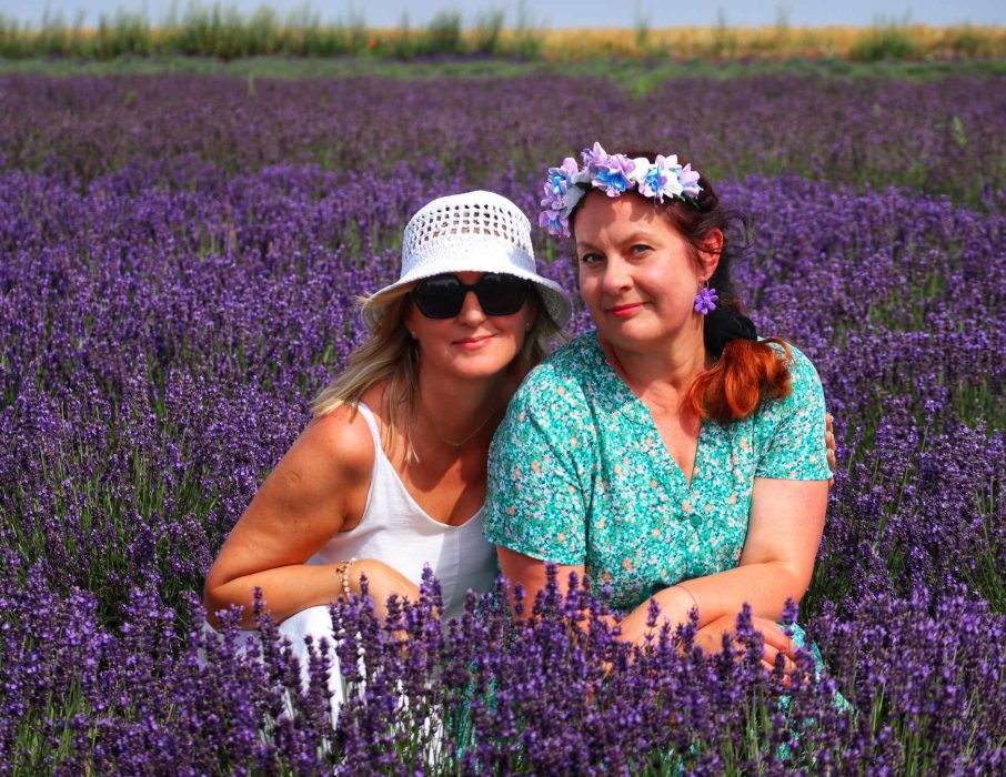 Monika Wojdyło (po prawej) w kwiecistej zielonej sukience i wianku siedzi obok uczestniczki warsztatów pośród rzędów fioletowej lawendy