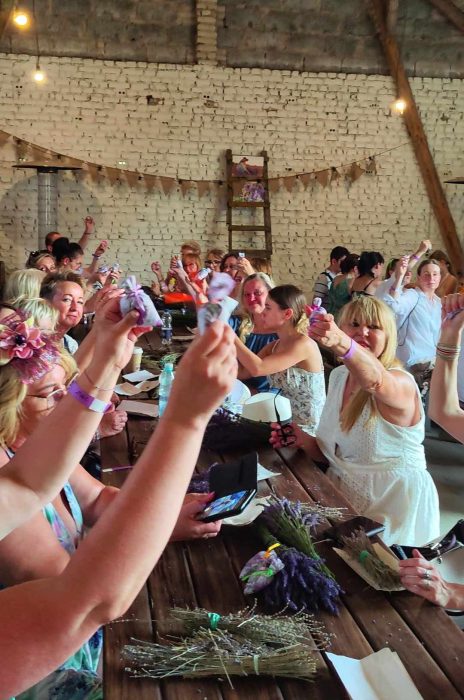 Grupa kobiet przy długim drewnianym stole w rustykalnej stodole unosi w górę własnoręcznie wykonane woreczki zapachowe z lawendą. Na stole leżą pęki świeżej i suszonej lawendy