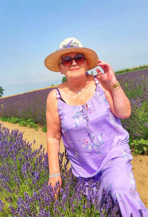 Kobieta w jasnofioletowej sukience w kwiaty i słomkowym kapeluszu z fioletową wstążką pozuje na tle rozległego pola lawendy pod bezchmurnym niebem