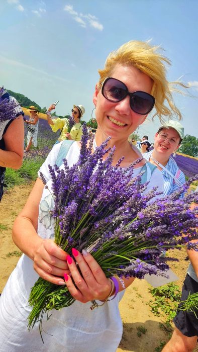 Uśmiechnięta kobieta w białej bluzce i okularach przeciwsłonecznych trzyma oburącz duży pęk świeżo ściętej, fioletowej lawendy na słonecznym polu