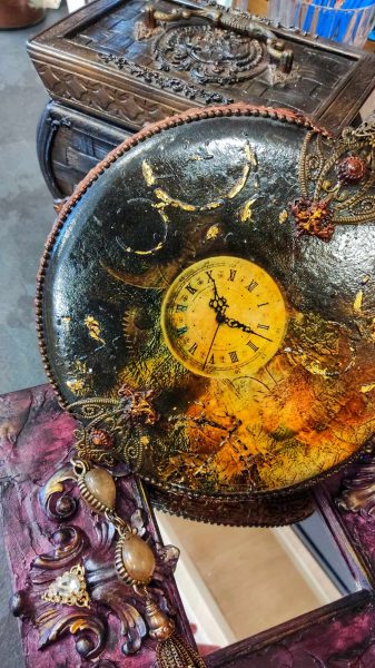 Okrągły zegar ścienny wykonany techniką decoupage, z tarczą w kolorze antycznego złota i czarną obudową z ozdobnymi, wypukłymi zdobieniami oraz złotymi przecierkami w stylu vintage