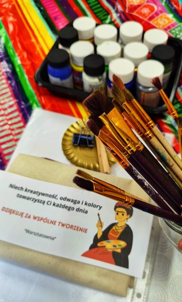 Zestaw materiałów malarskich do warsztatów Fridy Kahlo: farby akrylowe, pędzle różnych rozmiarów, instrukcja „Doświadcz wraz ze sobą kolory” i karta z artystką z Warsztatowni na kolorowym tle